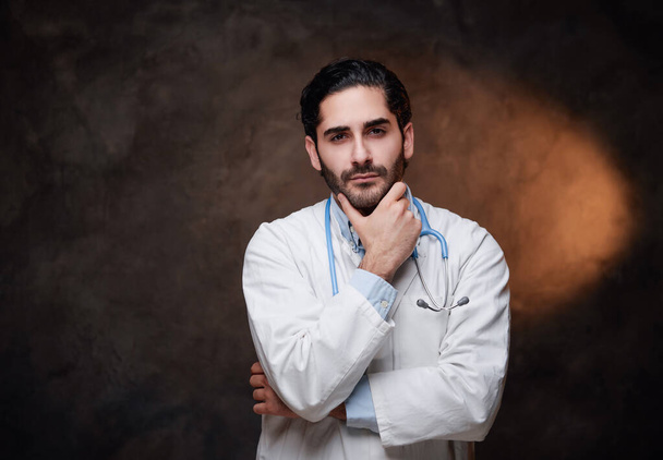 Zkušený lékař s plnovousem a bílým pláštěm v tmavém pozadí - Fotografie, Obrázek