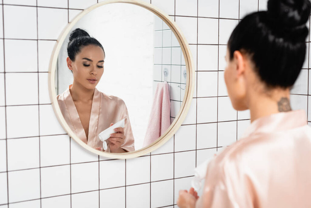 Mujer joven sosteniendo tubo con crema cosmética cerca del espejo en el baño  - Foto, Imagen