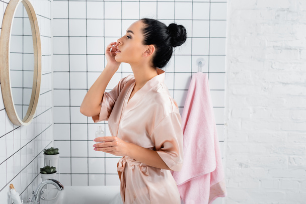 Mujer joven aplicando suero cosmético cerca del espejo en el baño  - Foto, Imagen