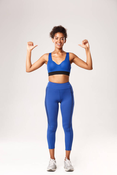 Pleine longueur de heureuse sportive afro-américaine confiante en tenue de sport bleue, se montrant avec un sourire fier et joyeux, montrant les progrès de l'entraînement, debout sur fond blanc - Photo, image