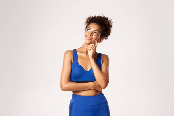 Stúdió lövés figyelmes mosolygós afro-amerikai fitness nő, visel kék sport ruhát, nézi a bal felső sarokban elégedett mosollyal, gondolkodás fehér háttér - Fotó, kép