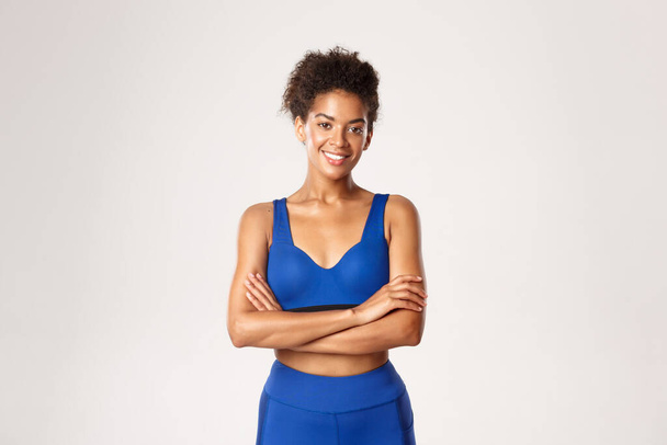 Atraktivní fit africko-americká fitness dívka v modrém sportovním oblečení, přes paže přes hrudník a s úsměvem, připraven na cvičení a cvičení, stojící na bílém pozadí - Fotografie, Obrázek