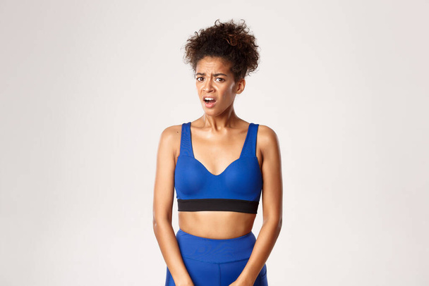 Közelkép a megdöbbent és elégedetlen afro-amerikai fitness lány, látszó frusztrált valami rossz, szemben fehér háttér - Fotó, kép