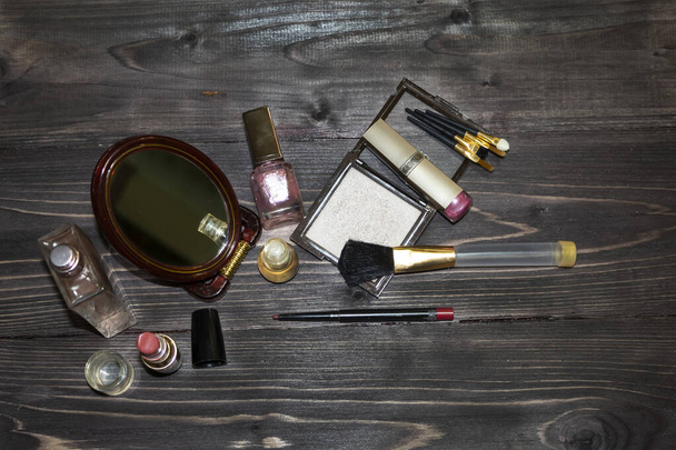 On a dark wooden background, there are a mirror, lipsticks, perfume, powder, pencil, brushes, nail polish. - Photo, Image
