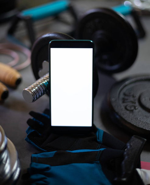 Мобильный телефон в окружении различных элементов для спортивных тренировок, таких как гантели, пластины для веса, сцепление для отжиманий, веревка для прыжков ... - Фото, изображение