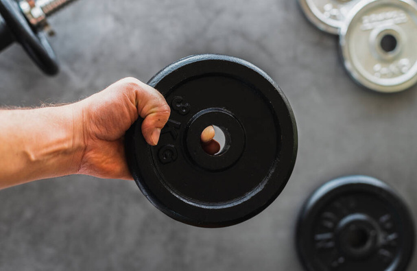 White person's hand holding a 3g iron weight plate over a background with dumbbells and plates of different weights - Фото, зображення