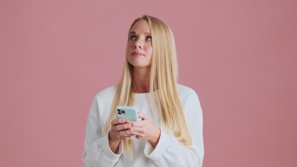 Een aantrekkelijke blonde vrouw is op zoek naar de zijkant terwijl met behulp van haar smartphone staan geïsoleerd over roze achtergrond in de studio - Video