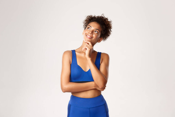 Photo studio d'une femme de fitness afro-américaine séduisante et réfléchie, portant des vêtements de sport bleus, regardant le coin supérieur gauche avec un sourire heureux, pensant à l'entraînement, fond blanc - Photo, image