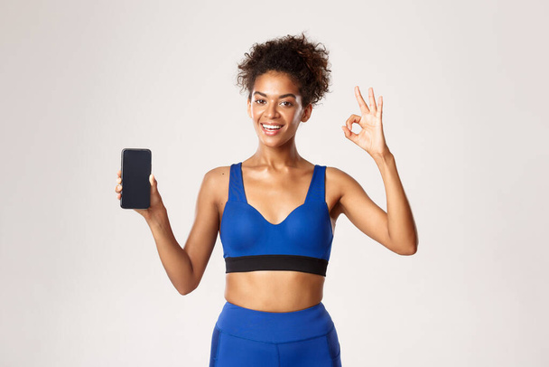 Concepto deportivo y tecnológico. Deportista afro-americana satisfecha en ropa azul de la aptitud, mostrando la pantalla del teléfono inteligente y la muestra aceptable, fondo blanco - Foto, Imagen