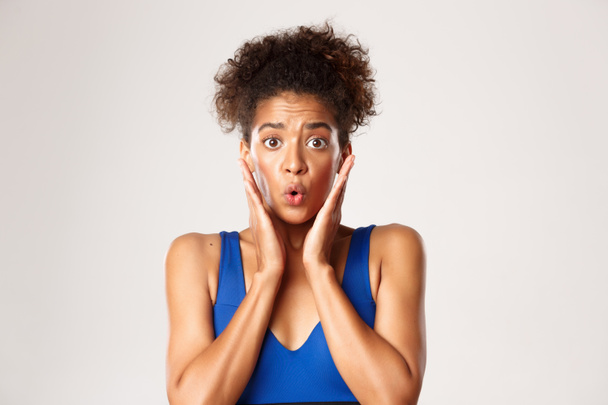 Concept of workout and sport. Close-up of alarmed and worried african-american female athlete, gasping and looking shocked, standing against white background - 写真・画像