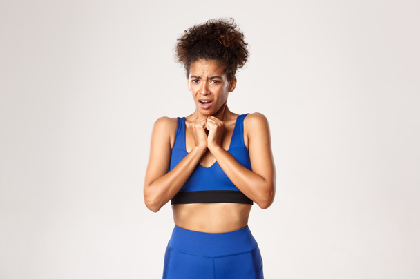 Waist-up of horrified african-american female athlete in blue sport oufit, grimacing and looking scared, standing against white background - Photo, Image