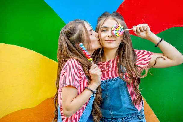 Schöne Teenager-Zwillingsschwestern in bunten Kleidern mit Lutschern auf buntem Hintergrund - Foto, Bild