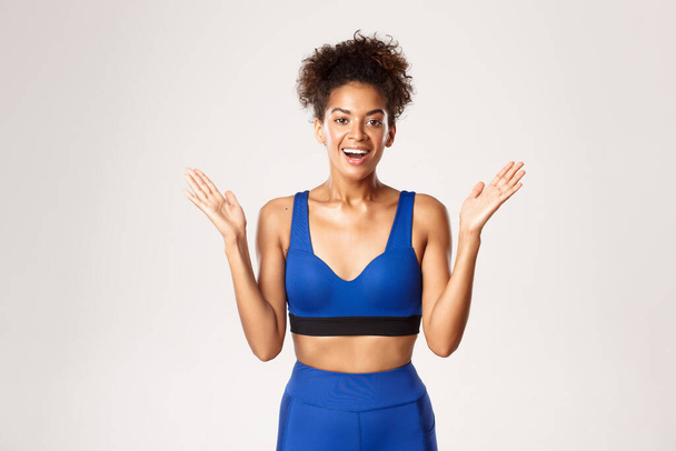 Waist-up van verrast en gelukkig, gezond Afrikaans-Amerikaanse fitness meisje in blauwe sport outfit, het verhogen van de handen omhoog blij en glimlachend, staande tegen een witte achtergrond - Foto, afbeelding