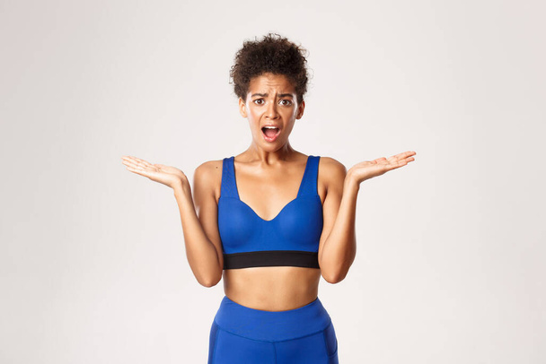 Περίμενε μια σοκαρισμένη και θορυβημένη Αφροαμερικανίδα αθλήτρια με μπλε στολή, να ουρλιάζει και να φαίνεται μπερδεμένη, να στέκεται πάνω σε λευκό φόντο - Φωτογραφία, εικόνα