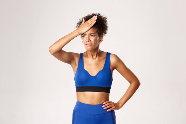 Estudio de la decepcionada mujer afroamericana fitness en traje deportivo azul, bofetada en la frente y mueca, de pie sobre fondo blanco - Foto, Imagen