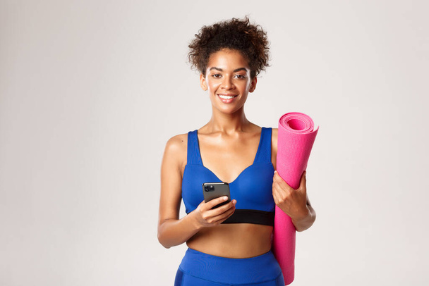 Waist-up shot of beautiful athletic woman in blue sports bra, smiling at camera, holding mobile phone and pink rubber mat for yoga or workout, white background - Photo, Image