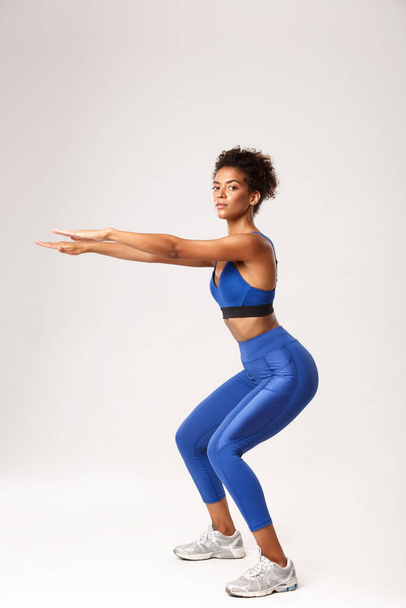 Полная длина красивой и здоровой африканско-американской женщины в синем спортивном костюме, протягивая руки для упражнений на корточках, глядя в камеру, беспокоясь на белом фоне - Фото, изображение
