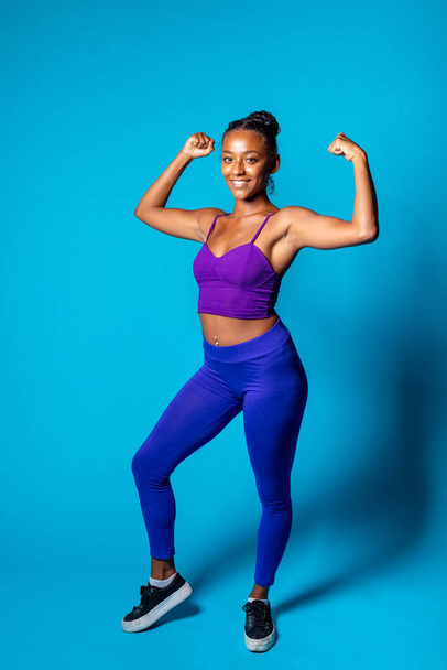 Fit jonge zwarte vrouw tonen spier op blauwe achtergrond - Geïsoleerde diverse zwarte vrouwelijke krachtige gebaar glimlachen - fitness, training, sport concept - Foto, afbeelding