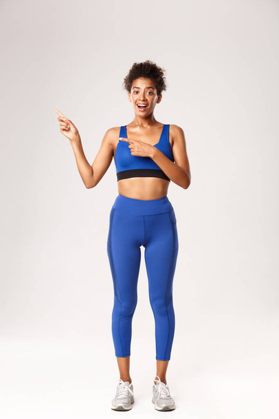 Teljes hosszában jóképű afro-amerikai sportos nő kék sportruházatban, ujjal mutogatva balra, hirdetést mutatva a sportról és az edzésről, fehér háttér felett állva - Fotó, kép