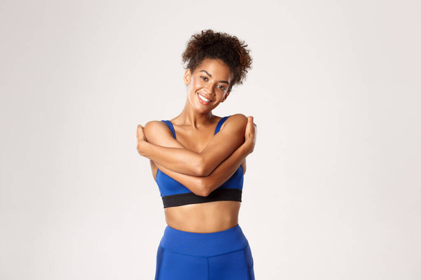 Concept van sport en training. Glimlachen gezonde Afrikaans-Amerikaanse vrouwelijke atleet, het dragen van blauwe sportkleding, knuffelen haar bazig en er gelukkig uitzien, staan tegen een witte achtergrond - Foto, afbeelding