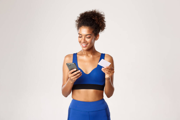 Forme attrayante femme de fitness afro-américaine, utilisant un téléphone portable et une carte de crédit, faisant du shopping en ligne, debout sur fond blanc - Photo, image