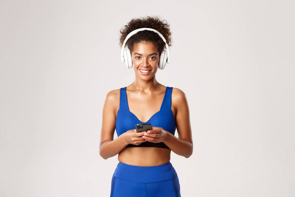 Joven deportista afroamericana en sujetador deportivo azul y leggings, sosteniendo smartphone, escuchando música en auriculares inalámbricos, ejercitándose sobre fondo blanco - Foto, imagen