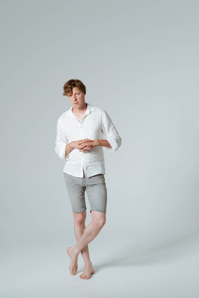 Pensive man standing full length folded arms and crossed legs. Thinking barefoot male model in white shirt demonstrates his emotions while posing on white background.  - Foto, Imagem