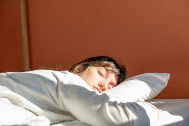 Νεαρή γυναίκα με ανοιχτόχρωμο δέρμα και καστανά μαλλιά κοιμάται στο κρεβάτι της μέχρι αργά με το φως του ήλιου να χτυπά το πρόσωπό της, λευκό κρεβάτι - Φωτογραφία, εικόνα