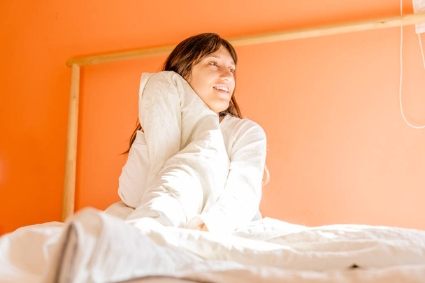Perfil da jovem mulher sentada na cama olhando para a janela em luz solar natural, mulher usa branco e sorri feliz - Foto, Imagem