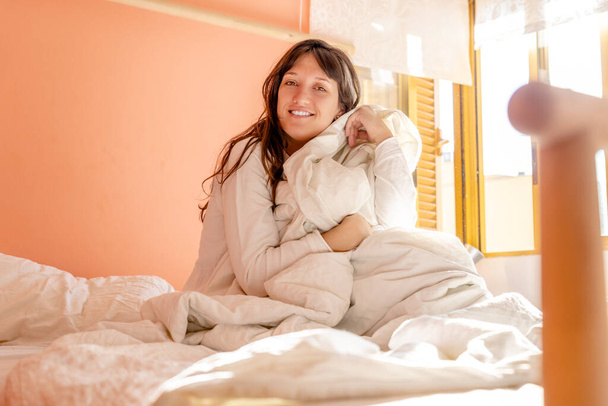 Mujer joven sentada en la cama vestida de blanco levantándose tarde con luz natural a través de la ventana, la mujer mira y sonríe a la cámara - Foto, imagen