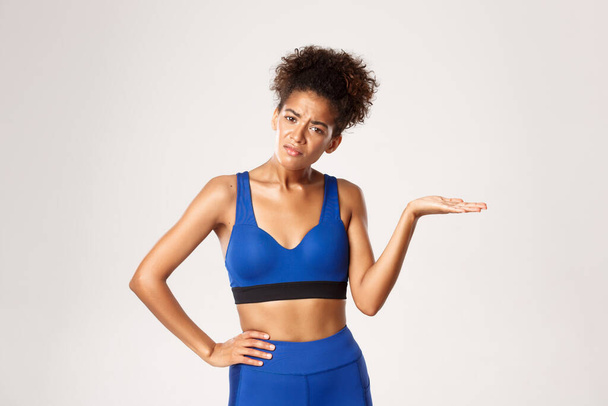 Studyjne ujęcie zdezorientowanej afro-amerykańskiej fitness kobiety, wzruszającej ramionami i wyglądającej nieświadomie, stojącej na białym tle - Zdjęcie, obraz