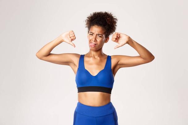 Tiro de estudio de atleta afroamericana decepcionada, vistiendo traje deportivo azul, quejándose como mostrando pulgares hacia abajo y la lengua pegada de aversión, fondo blanco - Foto, imagen