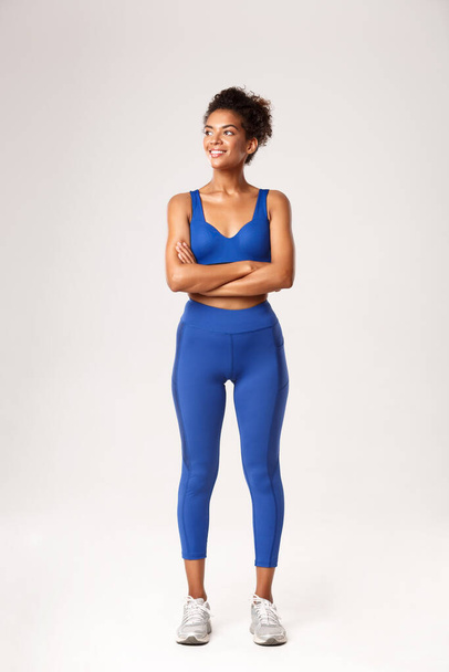 Full length of strong attractive african american sportswoman in sports bra and leggings, κοιτάζοντας αριστερά και χαμογελώντας σε κάτι, στέκεται πάνω από το λευκό φόντο, έτοιμος για προπόνηση - Φωτογραφία, εικόνα