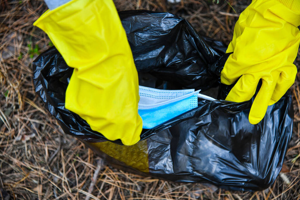 Umweltaktivist mit gelben Handschuhen zieht eine OP-Maske in einen Müllsack. Nach Ausbruch der Coronavirus-Pandemie begannen Mittel zum Schutz gegen COVID-19 die Umwelt zu verschmutzen. - Foto, Bild