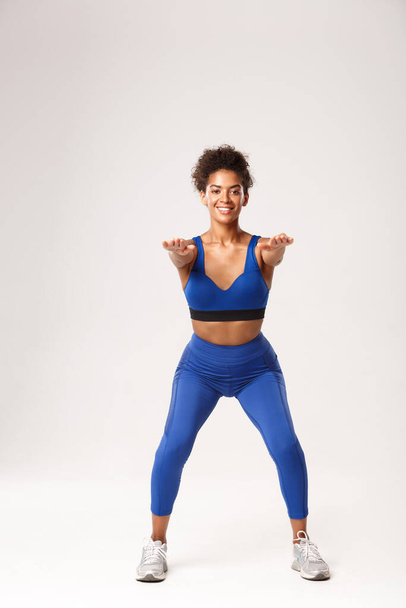 Pleine longueur de belle femme afro-américaine en tenue de sport, les mains tendues vers l'avant et faire des exercices de squats, souriant à la caméra, debout sur fond blanc - Photo, image
