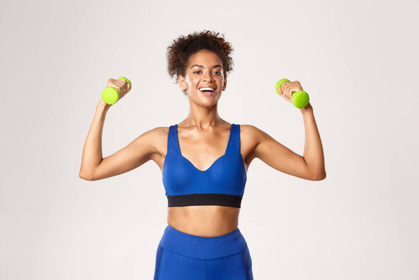 Concept de sport et d'entraînement. Forte et en forme femme de fitness afro-américaine vêtue de vêtements de gym bleus, faisant de l'exercice avec des haltères, debout sur fond blanc - Photo, image