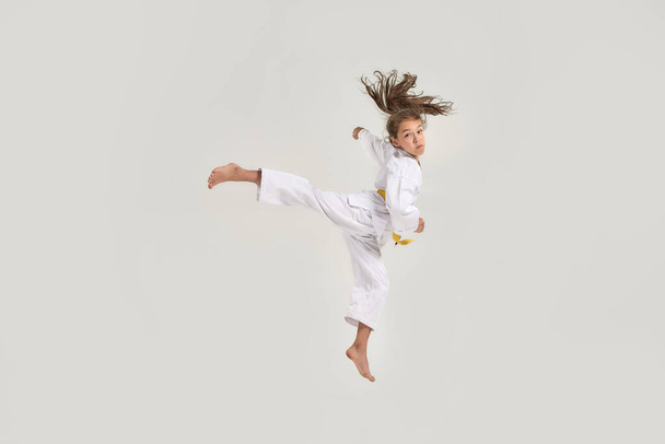 Full length shot of little karate girl in white kimono with a yellow sash exercising and fighting, doing martial arts, jumping isolated over white background - Φωτογραφία, εικόνα