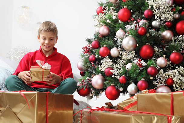Mutlu noeller ve mutlu tatiller, elinde çocuk hediye paketi ile süslenmiş Noel ağacının yanında, evde, oturma odasında.  - Fotoğraf, Görsel