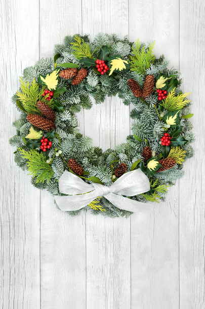 Winter solstice & Christmas  wreath with snow covered fir, cedar, holly, ivy, mistletoe, cedar, pine cones & white bow on rustic wood background. Composition for the xmas holiday season & New Year. - Photo, Image