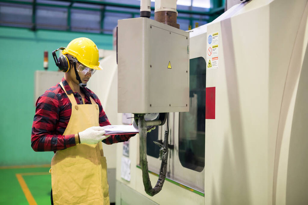再生シャツと安全ハード帽子を持つ工場労働者は、 CNC自動掘削機による金属鉄スペアパーツを生産するためにユーザーマニュアル紙をチェックします。高い技術を持つ重工業. - 写真・画像
