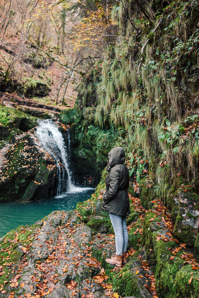 Girl in a jacket by the waterfall. Autumn or winter time in Europe. Woman enjoying cold weather and beauty of wild nature outdoors. Freedom Lifestyle. - Φωτογραφία, εικόνα