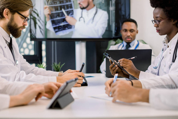 Cropped image of team of multiracial doctors using video screen projector during conference indoors, with their colleague discussing CT. African woman and Caucasian man making notes - Photo, Image