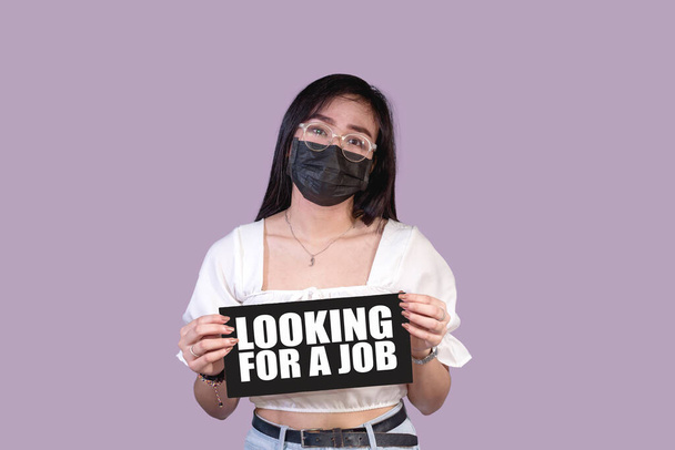 Una donna infelice che indossa una maschera facciale tiene una ricerca di un segno di lavoro. Licenziato, licenziato o disoccupato. - Foto, immagini