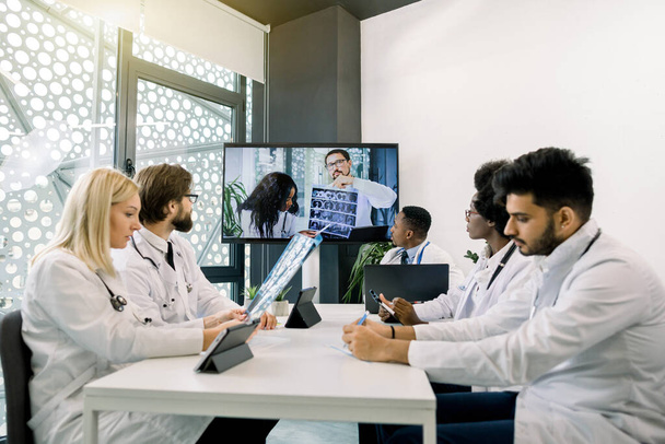 Front view of five multiracial confident doctors discussing treatment plan of patient while having video conference meeting with their Afrcian and Caucasian colleagues. Telemedicine, video chat - Fotografie, Obrázek