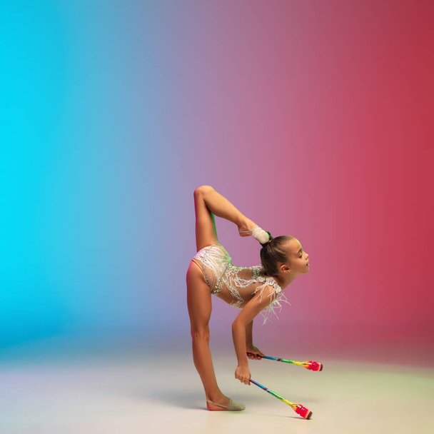 Little caucasian girl, rhytmic gymnast training, performing isolated on gradient blue-red studio background in neon - Foto, imagen