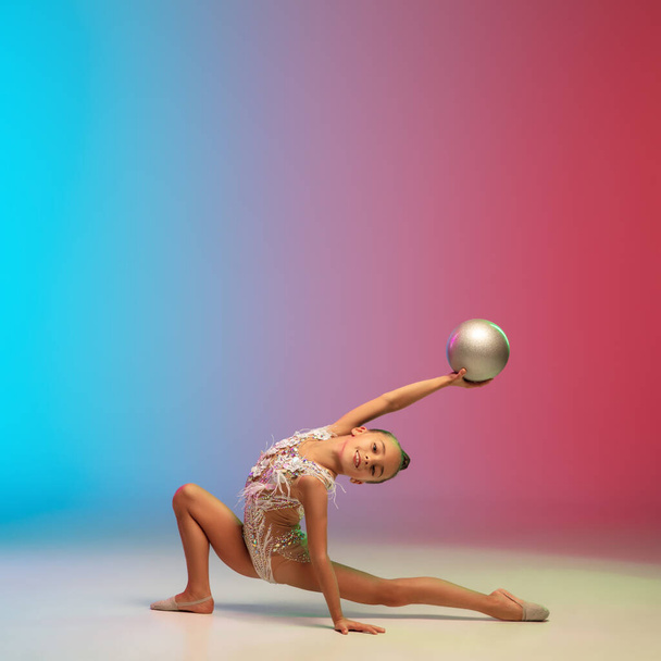 Mała kaukaska dziewczyna, gimnastyka rytmiczna trening, wykonywania izolowane na gradientowym tle niebiesko-czerwony studio w neonie - Zdjęcie, obraz
