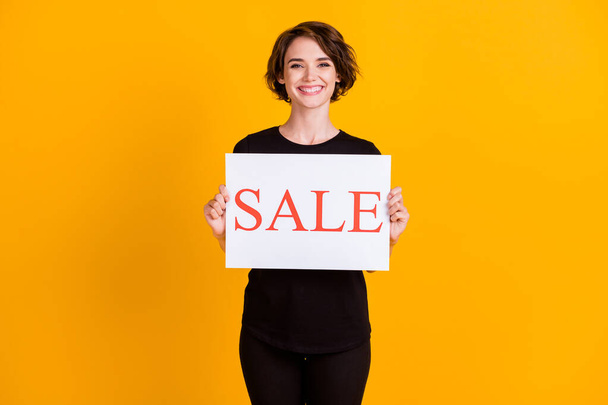 Portrait of attractive cheerful girl holding in hand paper placard sale advert isolated over bright yellow color background - Foto, imagen