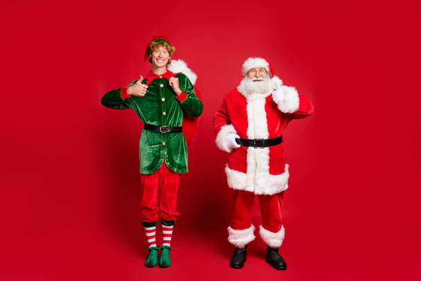 Full length photo of two people santa elf hold big bag raise thumb up wear x-mas costume cap glasses boots isolated red color background - Foto, immagini