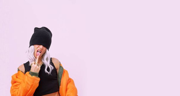 Blonde model in fashion urban street look. Trendy orange bomber jacket and stylish beanie cap. Middle finger provocation. Fall winter seasons lookbook - Photo, image