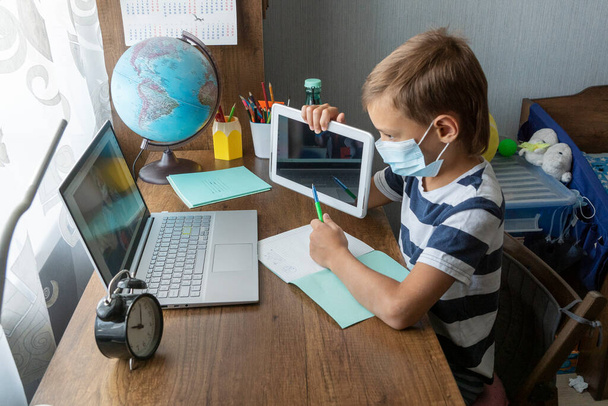 scolaro in maschera viso medico mostra compiti a casa sul suo tablet per insegnante sullo schermo del computer portatile durante la lezione online. Istruzione a distanza durante l'isolamento. - Foto, immagini